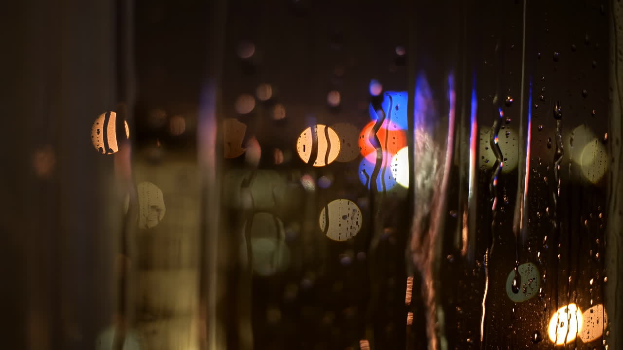 雨粒がついた窓、ぼやけた都会の光と温かいボケの輝き、映画やロマンスにぴったりの親密な夜の雰囲気。