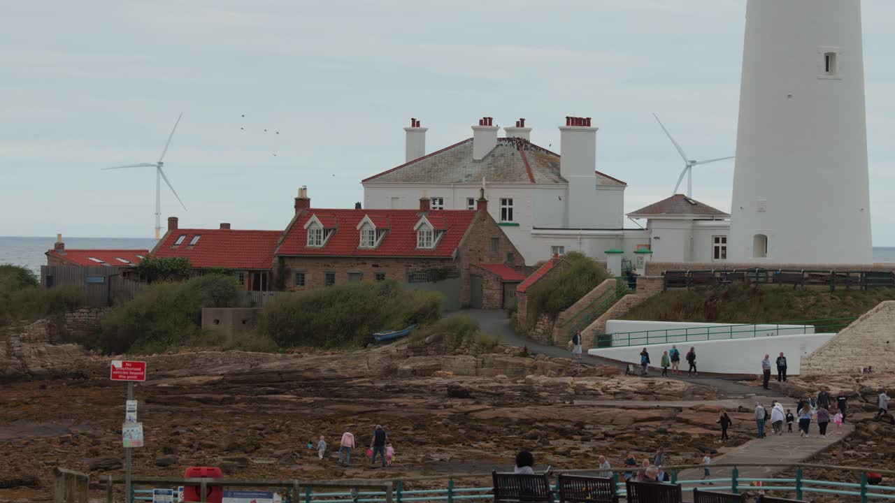 Groups walk along rocky coast by tall white lighthouse, cottages, and distant wind turbines