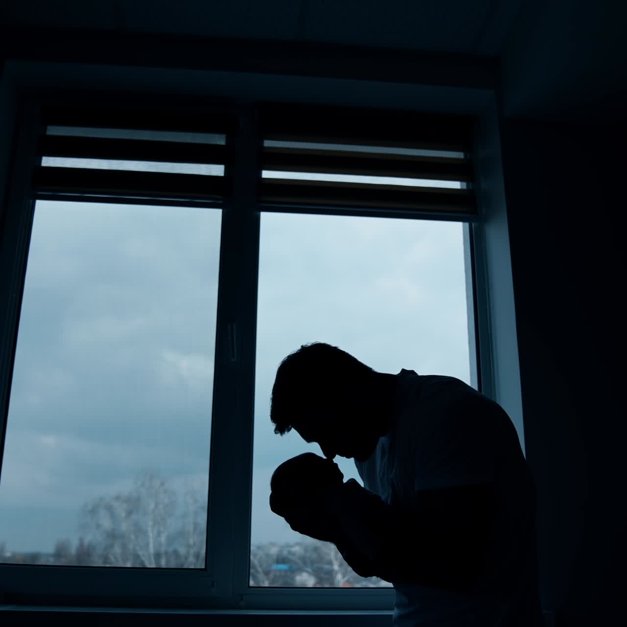 Big strong man's silhouette with a baby in hands. Father holding his newborn standing in the dark room in front of the window