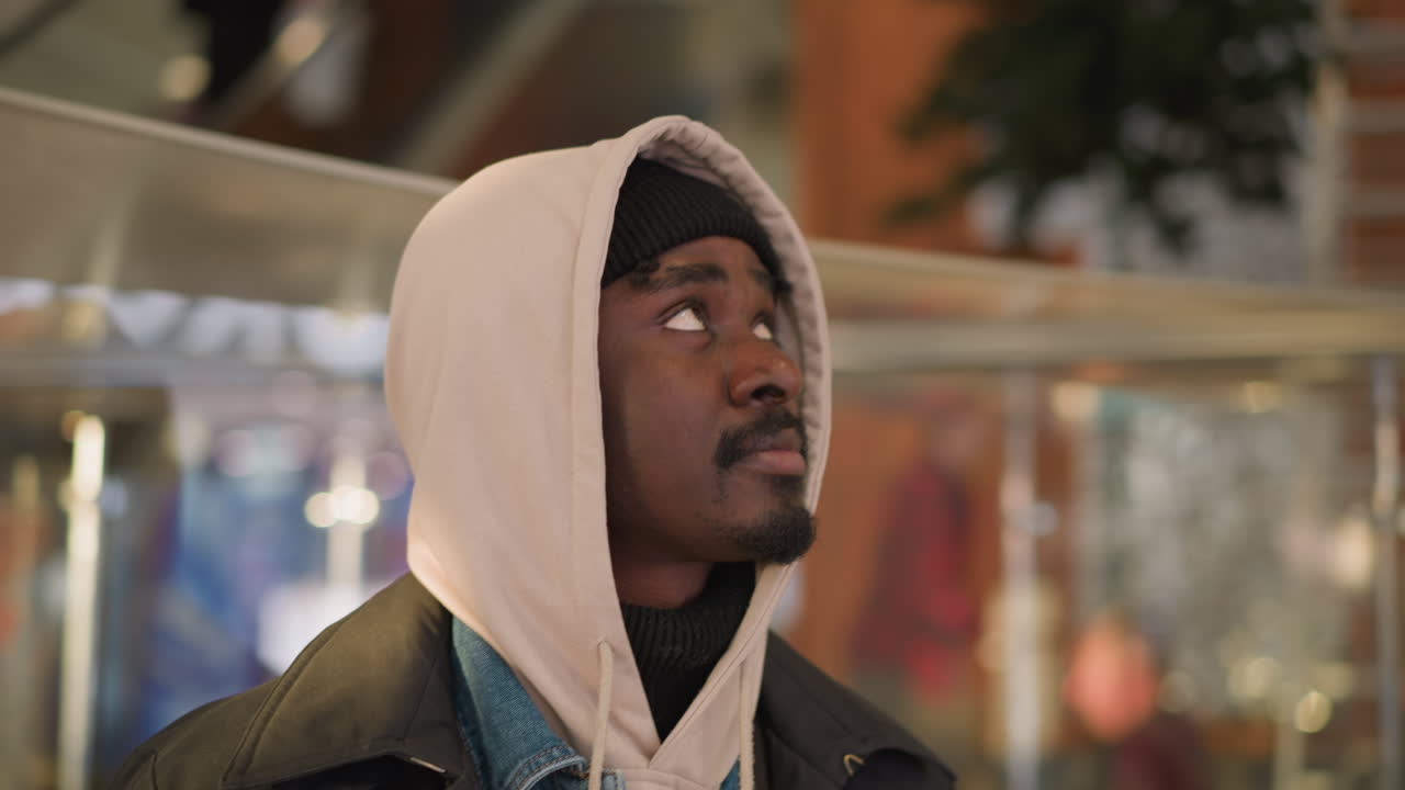 Young black man wearing beige hoodie and black beanie looking upward inside modern glass atrium with blurred lights and stainless steel railings exhibiting thoughtful introspection and urban calm vibe