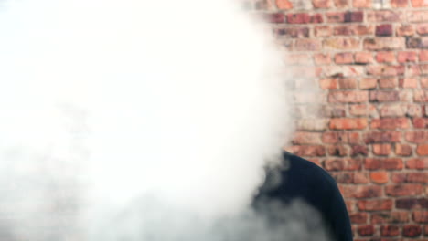 portrait of a man vaping indoors in front of a brick wall with cloud of smoke