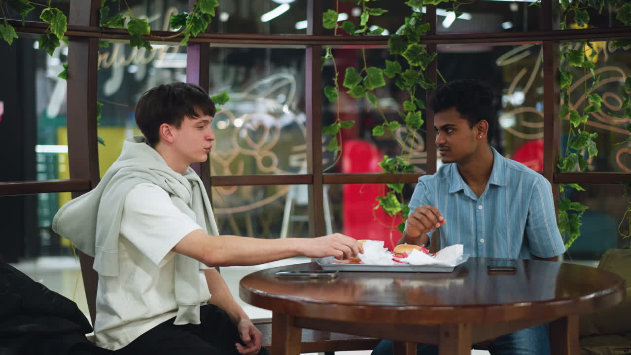 Two men facing each other discussing while grabbing fries from tray, candid interaction in casual dining space, expressions engaged, close framing shows hands reaching for crispy potato sticks