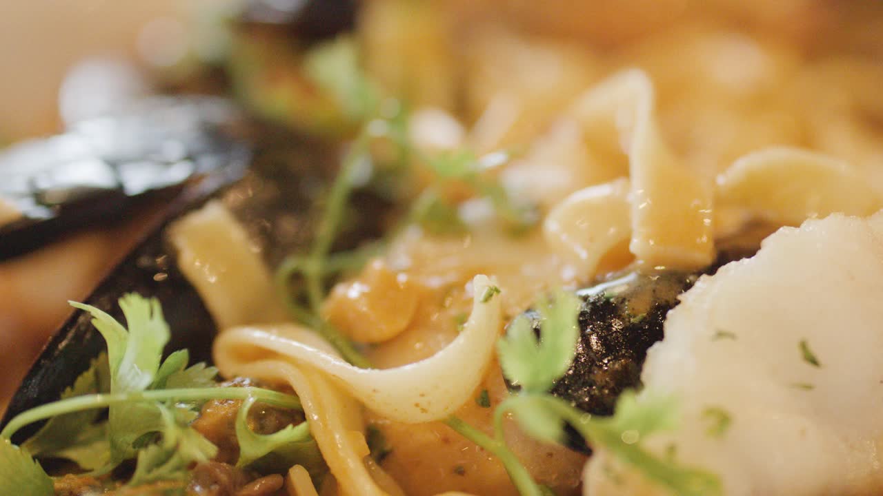 Macro view of assorted shellfish, scallops, and noodles in a hot seafood chowder, garnished with fresh herbs under warm, soft lighting. Subtle camera movement enhances texture