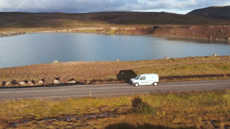 Profile-shot-of-white-car-traveling-on-rural-road-along-lake-with-bright-sunlight,-dusk