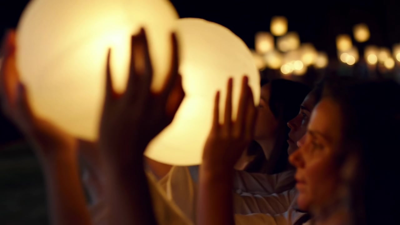 Outdoors after dark, people stand in line holding illuminated paper lanterns close to faces, eyes lowered in quiet focus, creating gentle warmth, soft light, and shared contemplative mood