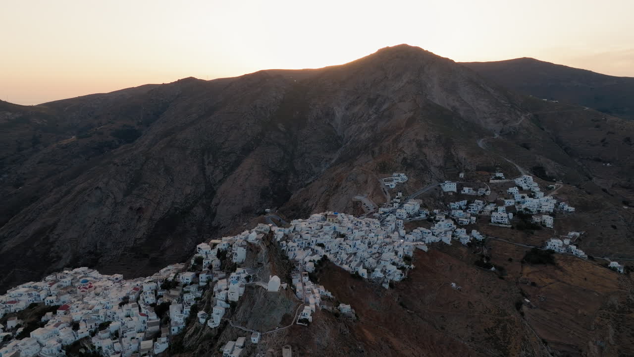 Serifos tepeleri ve Chora şapeli üzerinde puslu gün batımı, drone silüeti ve karanlık araziyi görüntülüyor.