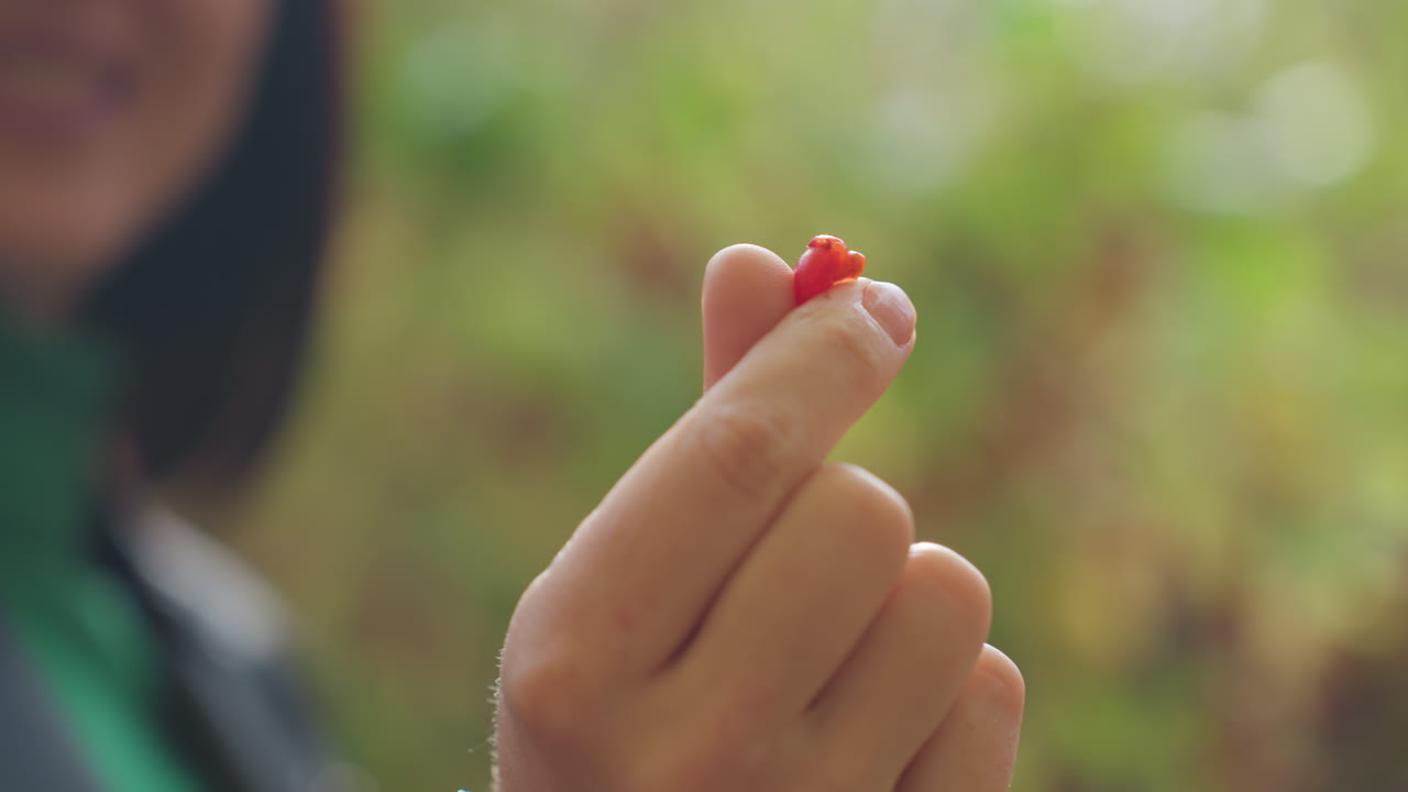 mujer asiática con chaqueta de cuero que sostiene una baya entre el pulgar y el índice, ligera sonrisa, manicura visible, fondo de jardín al aire libre con bokeh verde, luz suave y cálida, ambiente de músico de rock, íntimo