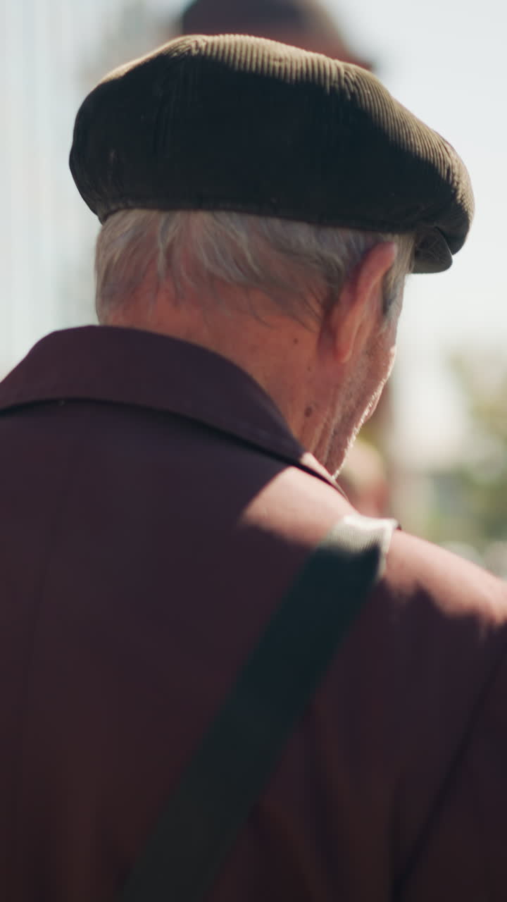 Uomo anziano bianco visto da dietro con berretto e tracolla. L'inquadratura ravvicinata cattura il cappotto texturizzato, i capelli grigi, le rughe e il morbido sfondo bokeh della città, la calda luce del sole pomeridiano e la tranquilla atmosfera nostalgica