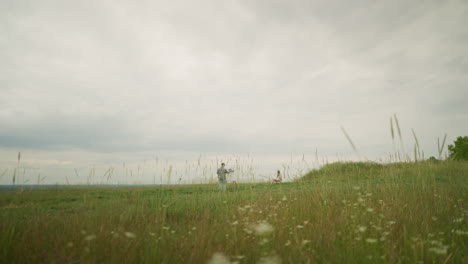 a composer, wearing a hat and checkered shirt, is intensely focused on painting on an easel in a vibrant grass field under a cloudy sky. a woman in a hat and white gown sits calmly in a chair