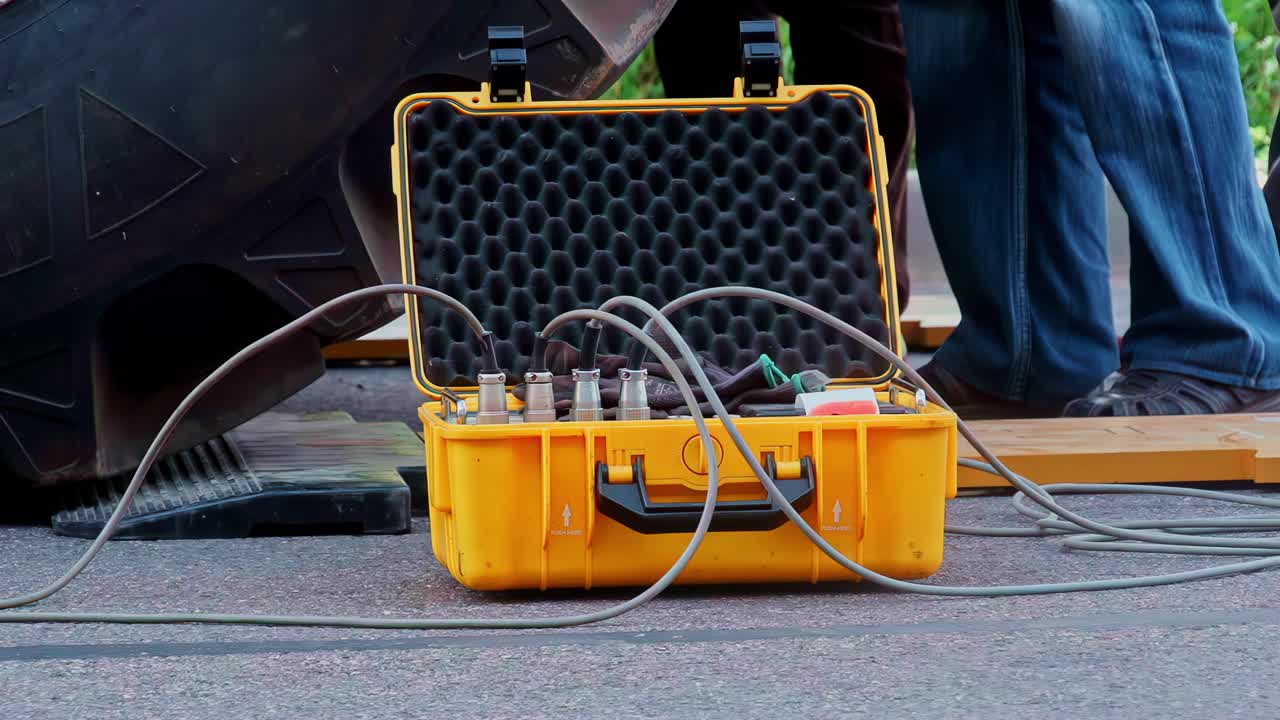 Statisk bild av en gul robust utrustningsväska med anslutna kablar och sensorkontakter på marken vid en byggarbetsplats, bärbar mätutrustning och fälttestsats bredvid tunga maskiner.