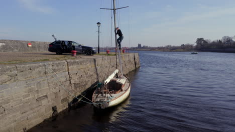 sailboat moored along pier, man works on deck climbing up mast under calm sunlight after storm, tracking tilt up