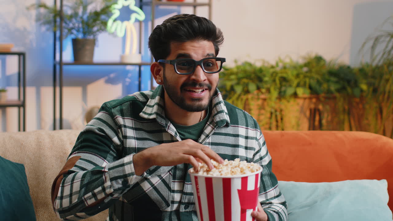 Indian man sits on sofa eating popcorn and watching interesting tv serial sport game online at home
