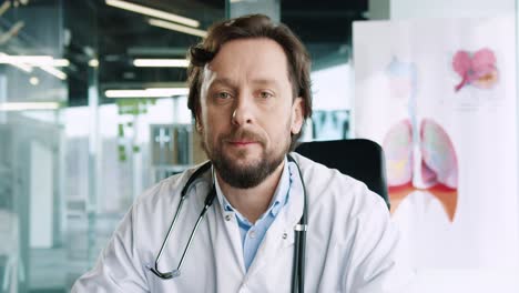 Close-up-view-of-caucasian-senior-male-doctor-with-stethoscope-sitting-at-desk-looking-at-camera-and-smiling-in-hospital