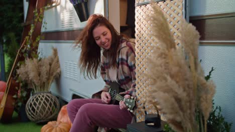 Happy-brunette-girl-in-a-checkered-shirt-and-pink-jeans-plays-a-black-ukulele-near-her-trailer-outside-the-city-in-the-summer-during-a-picnic.-Yellow-sprigs-of-reeds-in-a-vase-and-other-decorations-of-the-trailer-during-their-day-off-outside-the-city-in-the-summer-on-a-picnic