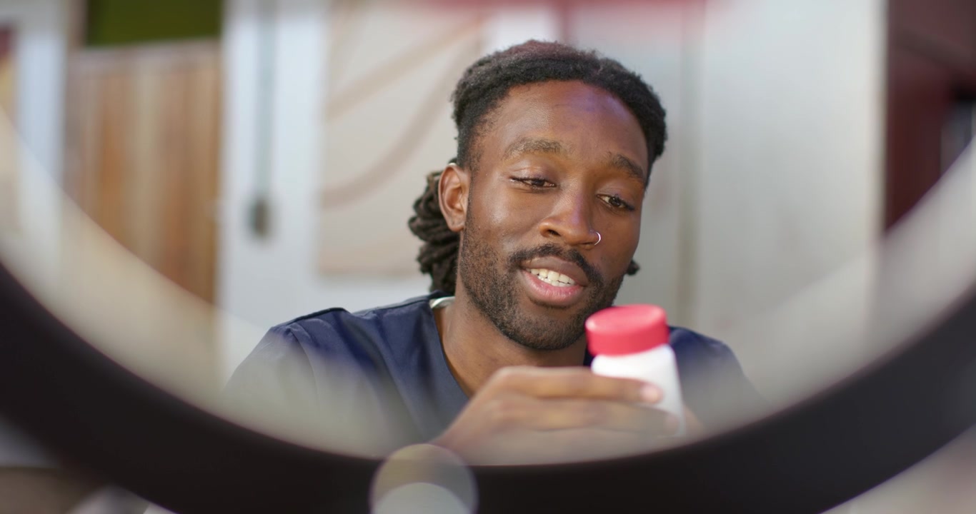 Messa a fuoco del telefono che si sposta, mentre un uomo africano adulto si allunga, mostrando una bottiglia bianca con tappo rosso.