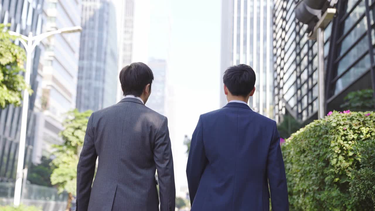 市街地の中心部の路上で話しながら歩く2人のアジア人ビジネスマンの後ろ姿。