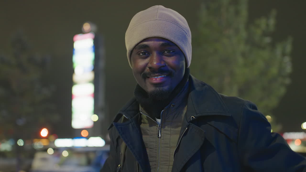 Happy man wearing warm coat and hat smiles directly at camera during cold winter night outdoors with blurred city lights glowing in background