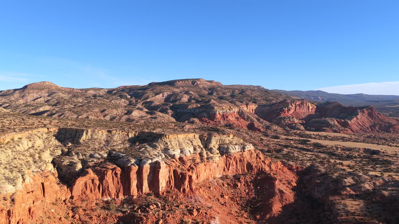 Una dinamica ripresa con drone avanza sulle aguzze e verticali formazioni di arenaria di Abiquiu verso il distante orizzonte montuoso. Questa transizione cinematografica cattura la bellezza aspra del New Mexico.