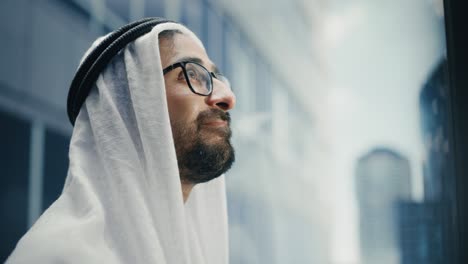 portrait of a successful happy muslim businessman in traditional white kandura wearing glasses riding glass elevator to office in modern business center. saudi, emirati, arab businessman concept.