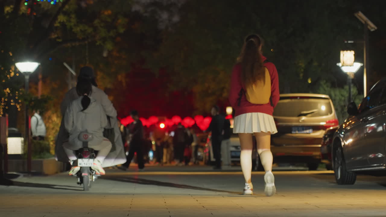 Giovane donna che cammina verso una folla con lanterne su una strada poco illuminata mentre passa uno scooter, bagliore di lanterne rosse, una figura con zaino, in gonna bianca e giacca rossa, si avvicina a un festival vivace, atmosfera notturna.