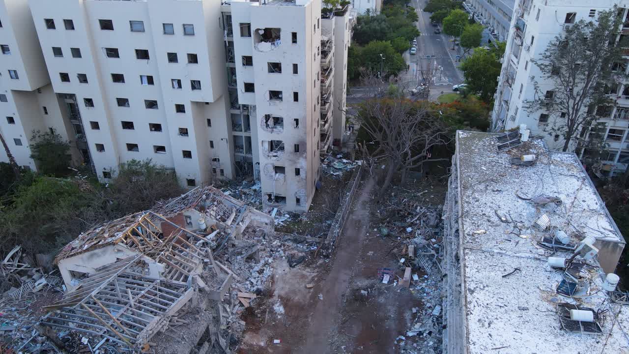 Drone pans over damaged buildings in Tel Aviv, showing debris, shattered windows, and an uprooted tree.
