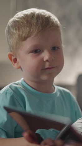 chubby blond toddler watches cartoon with mom and sister on tablet. curious little boy in blue shirt talks looking at screen of gadget closeup