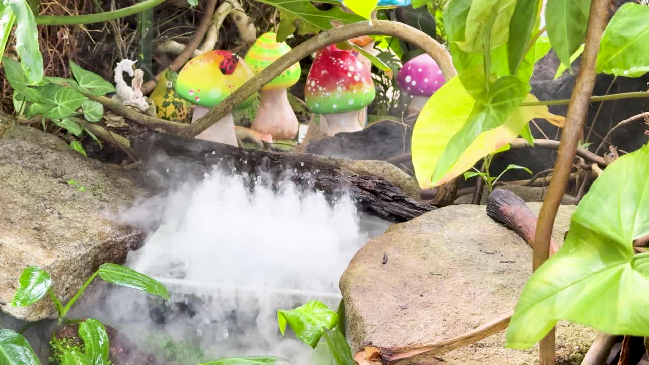 Artificial mist flows from whimsical garden fountain with colorful mushrooms, surrounded by vibrant foliage, daylight