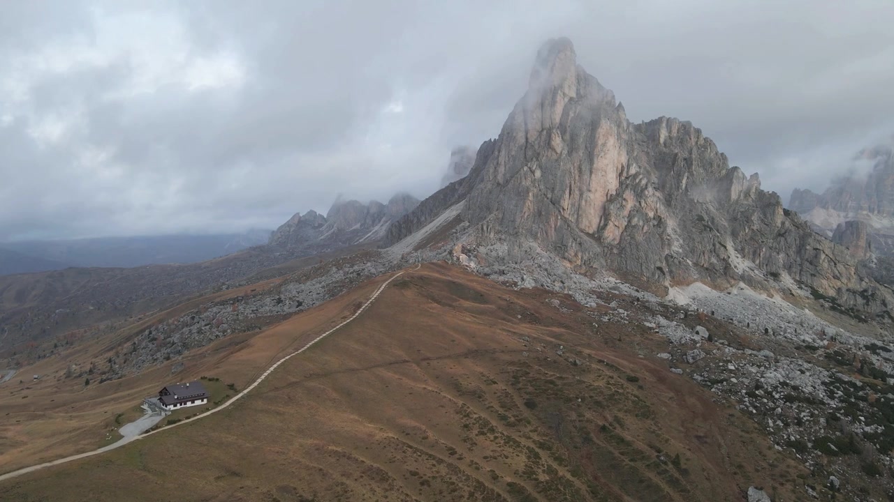 Vista maestosa del Passo Giau in Alto Adige, Italia, con cime imponenti, un rifugio alpino e terreno roccioso sotto un cielo nuvoloso. La scena serena ma drammatica esalta la bellezza alpina.