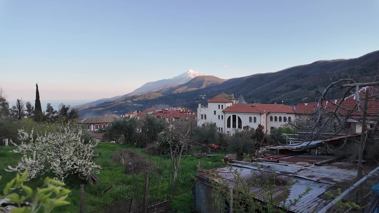 Sakin bir köyde oturan insanlar, dağların üzerindeki gün batımını izliyor. Köyde kırmızı çatılı birçok ev ve yakınlarda ağaçlar bulunuyor.