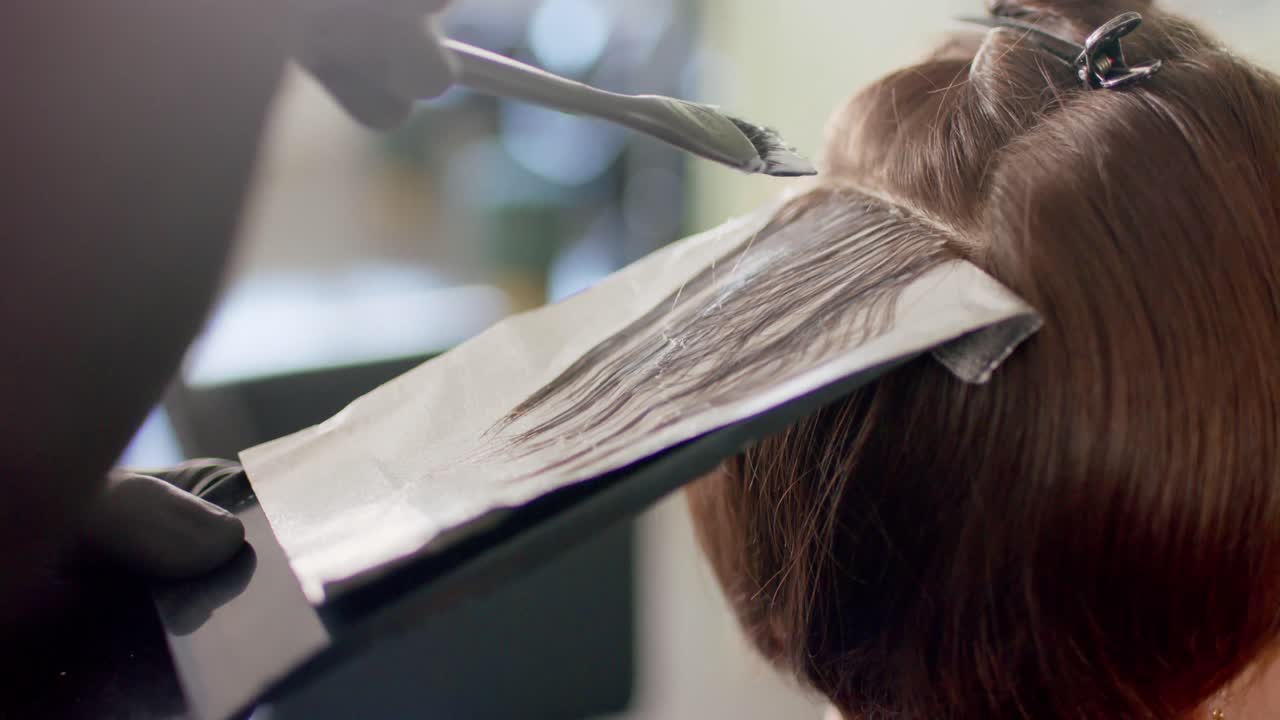 Close up of hairdresser highlighting client's hair with brush and foil at salon, in slow motion