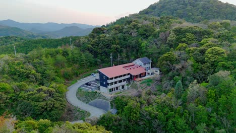 a slow aerial pan reveals a secluded lodge nestled in a lush green forest. the shot showcases the building, a winding driveway, and the vast, scenic mountain range in the background
