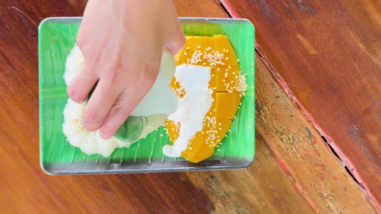 A hand pours coconut sauce over heart-shaped sticky rice and sliced mango on a green plate.