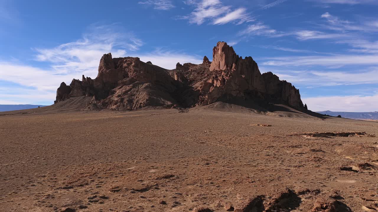 Un drone si avvicina a un avamposto montano accidentato, evidenziando le cime rocciose frastagliate e l'ambiente selvaggio e desolato di una landa desertica ristretta o post-apocalittica.