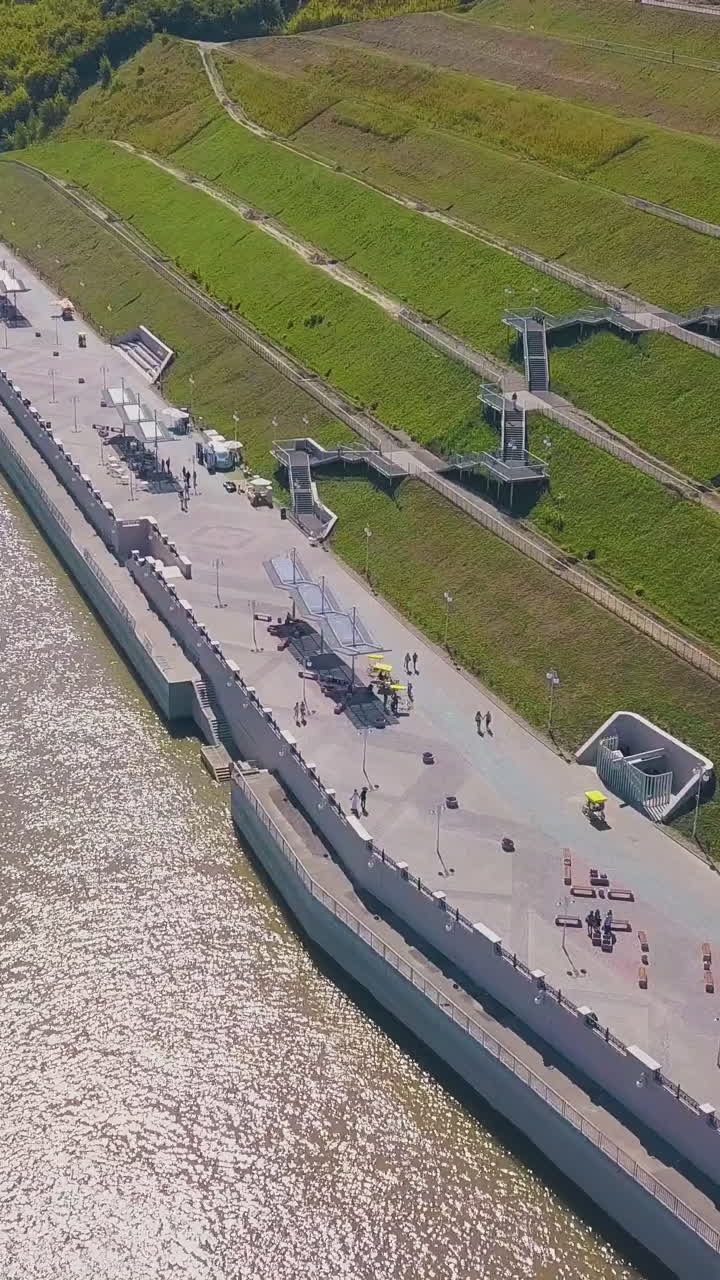 distant newlywed couple walks along waterfront near calm river and cascade hill on sunny day aerial view