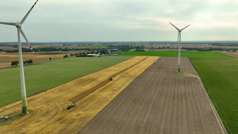 Luftaufnahme-Von-Windturbinen,-Die-Hoch-Auf-Einem-Großen-Landwirtschaftlichen-Feld-Stehen,-Mit-Einem-Traktor,-Der-Auf-Dem-Abgeernteten-Weizenfeld-Sichtbar-Ist