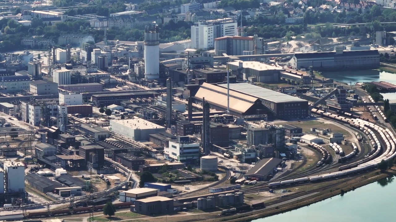 Panoramic view of a large steel manufacturing complex with blast furnaces and chimneys. The industrial zone is located near a river and forest