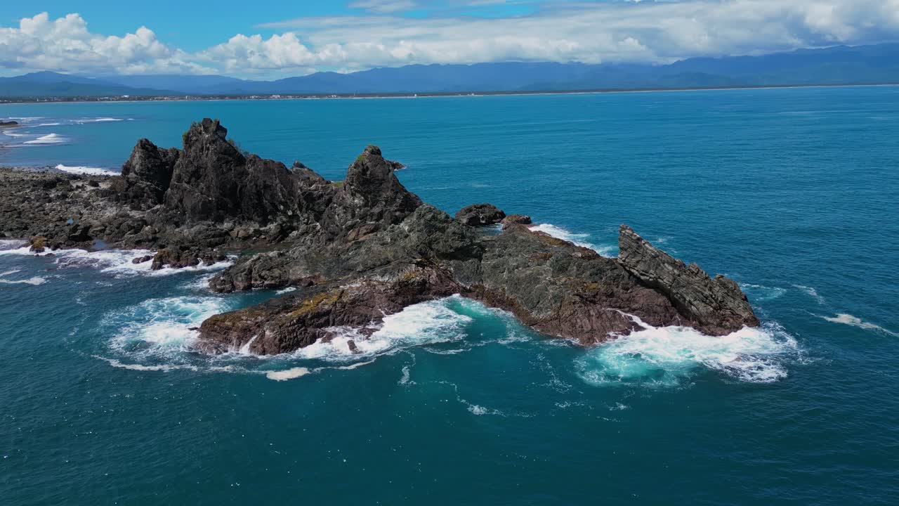 A dramatic drone shot captures the small rocky mountains rising from the shore, with the ocean waves crashing gently at their base.