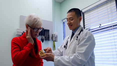 Young-Asian-male-doctor-and-senior-patient-discussing-over-ear-model-at-clinic-4k