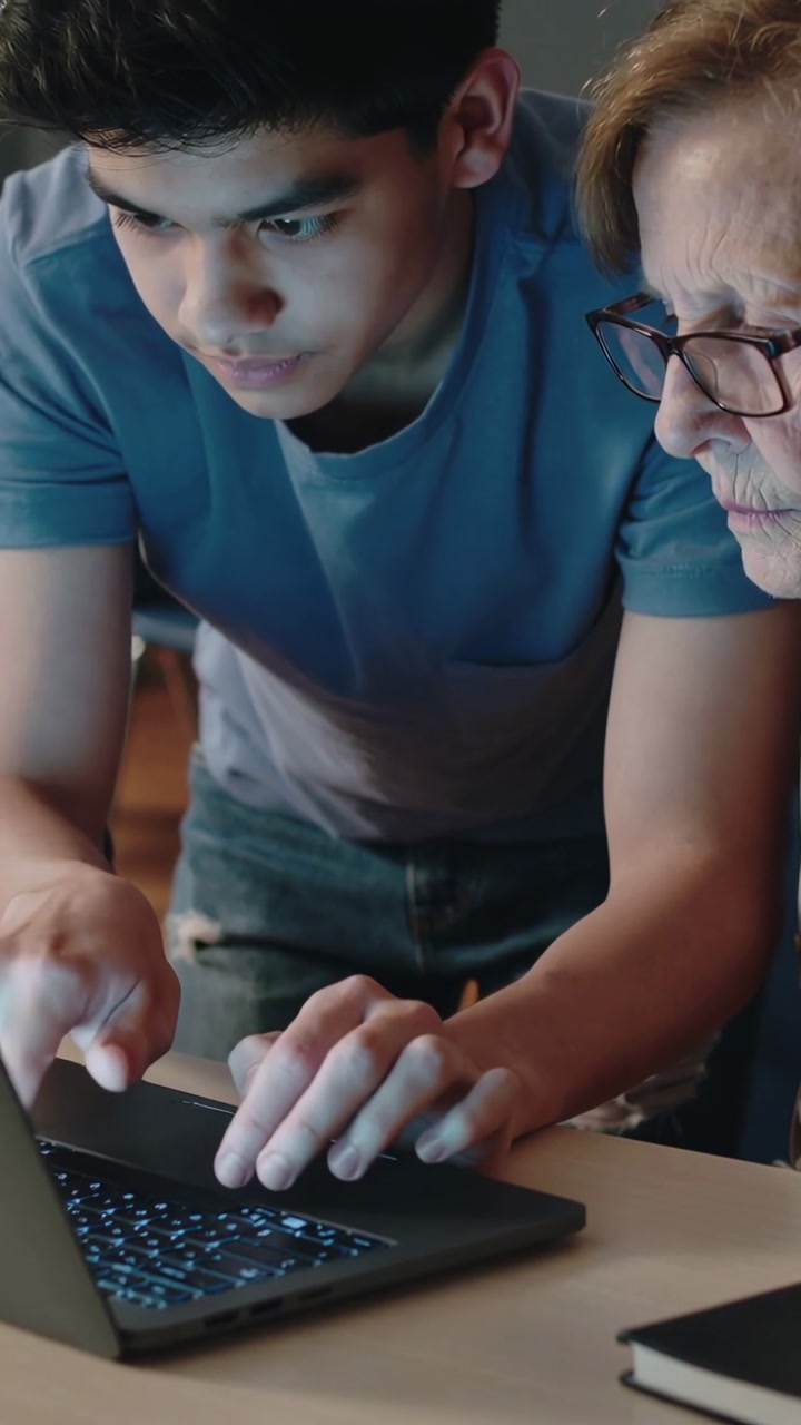 Vertical video: Leaning teenager typing and pointing at laptop at home, guiding Senior with glasses