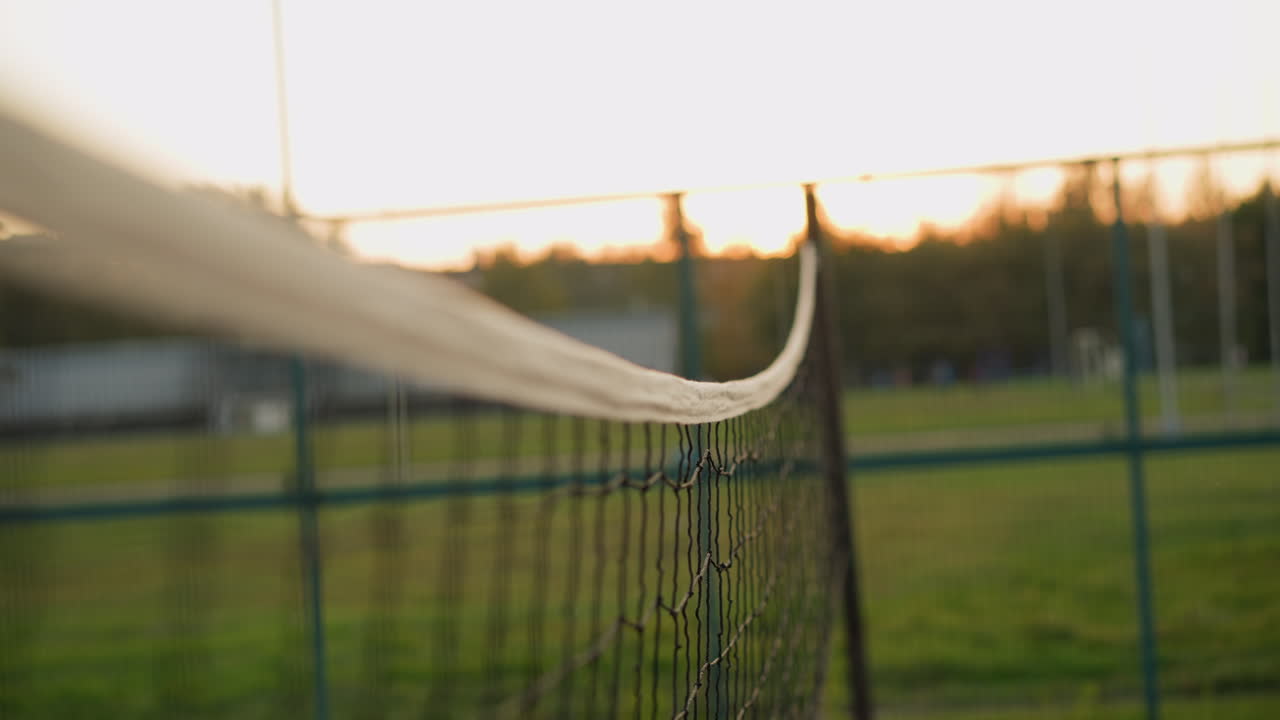 볼리볼 네트의 클로즈업, 선수들의 손이 공을 때리기 위해 손을 는 모습, 배경의 야외 코트, 해가 지는 동안의 볼리볼 경기의 활기찬 순간