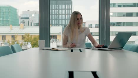 young business woman in conference room with papers. elegant blond woman sitting at long table in modern hall of office with laptop and writing on papers while working