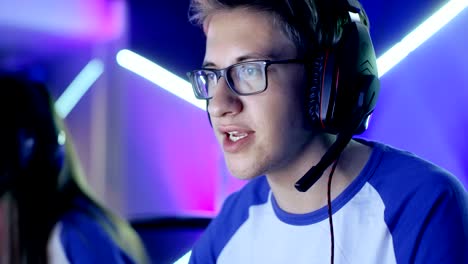 teenage boy gamer plays in competitive video game on a esports tournament/ internet cafe. he wears glasses and headphones and speaks into microphone.