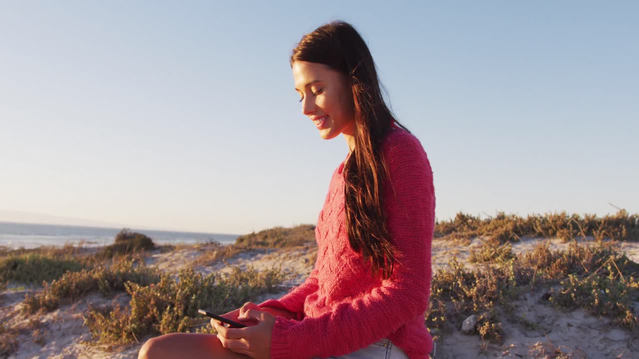 Glad kaukasisk kvinde sidder på stranden ved havet og bruger smartphone.