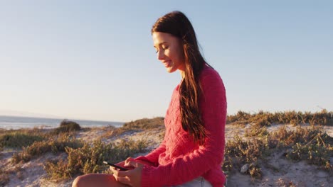 Feliz-Mujer-Caucásica-Sentada-En-La-Playa-Junto-Al-Mar-Usando-Un-Teléfono-Inteligente