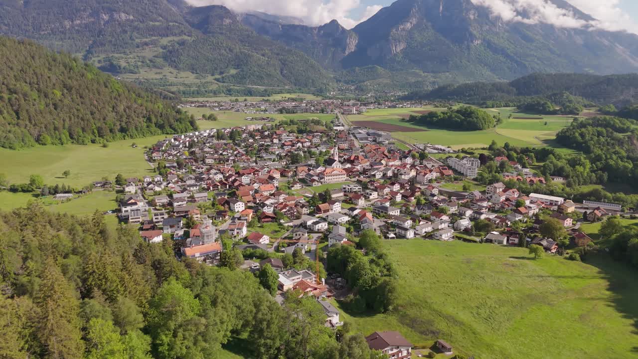 Forest trees on hill of mountain and historic town in distance. Sunny day with alps in Back. Aerial wide shot. Houses and homes surrounded by green lawn field.