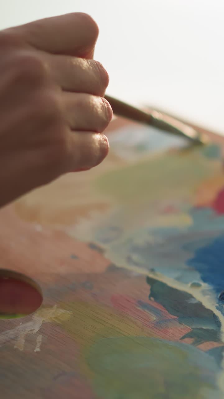 una vista de cerca de una mano femenina mezclando delicadamente pinturas en una paleta con un pequeño pincel. la luz suave mejora el proceso artístico, capturando el movimiento y la sutileza de la mezcla de colores