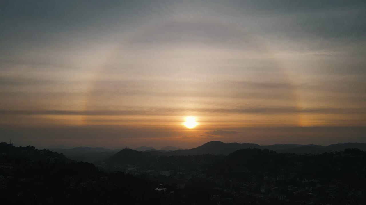 Beautiful setting sun with a rainbow circle between the thin veil clouds above the silhouettes of the mountains from the nature of Sri Lanka near Kandy. High level drone shot
