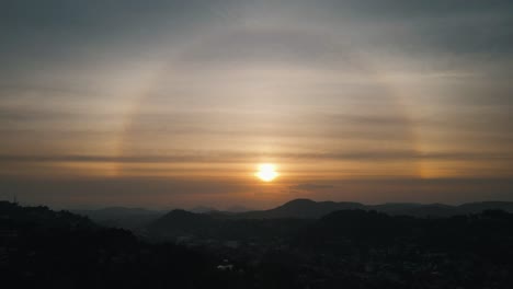 beautiful setting sun with a rainbow circle between the thin veil clouds above the silhouettes of the mountains from the nature of sri lanka near kandy. high level drone shot