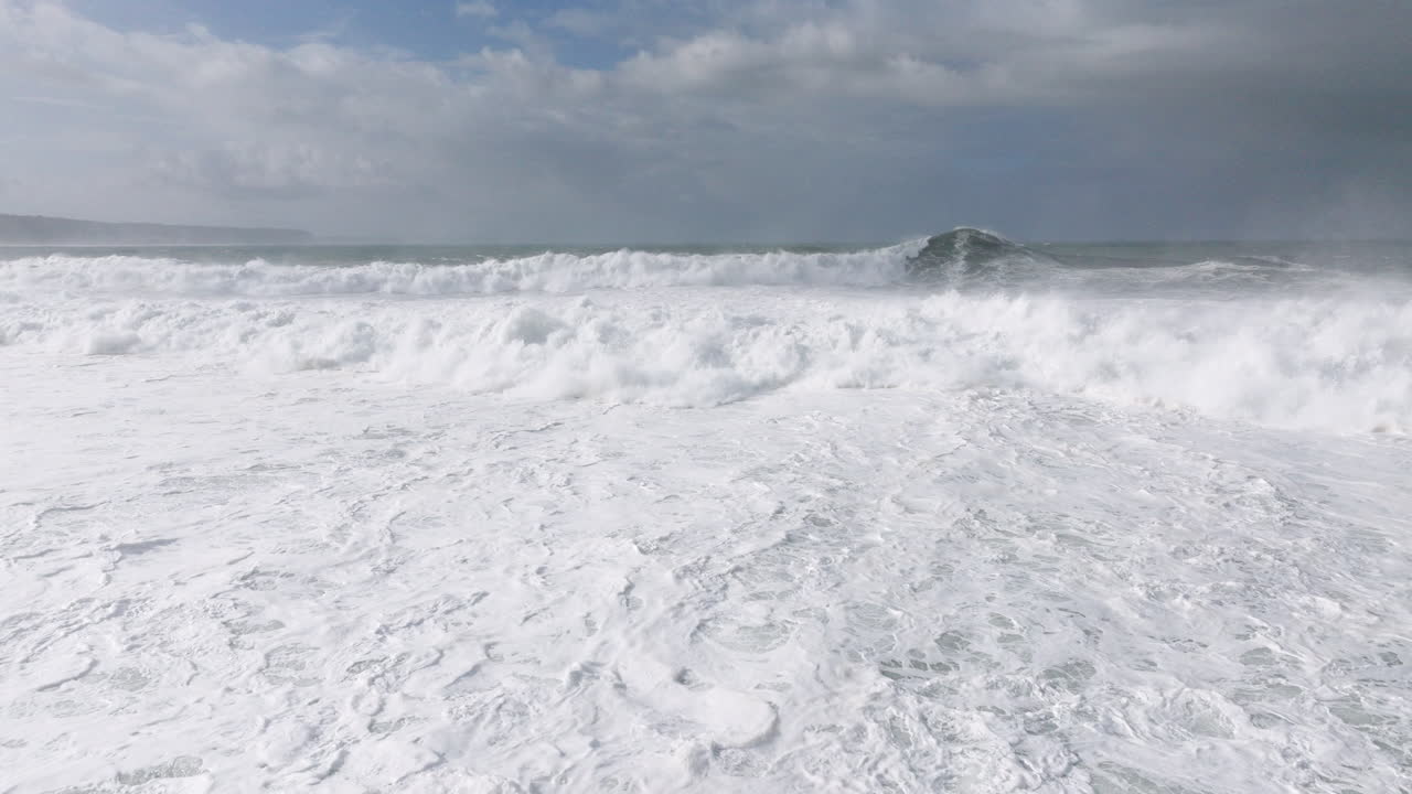 Aerial drone shot of big waves coming into shore on a day with giant waves in Nazaré, Portugal, Europe. Drone flying low and towards white surf and breaking wave line at Nazare