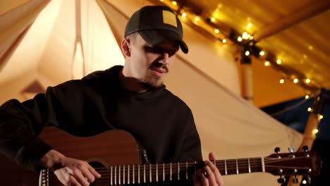 man plays the guitar and sings songs in a tent with light bulbs